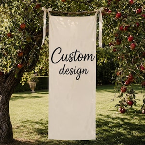 Pode incluir: Uma faixa retangular de tecido na cor off-white pendurada no galho de uma árvore por duas fitas claras. A faixa apresenta o texto preto Custom design impresso em uma fonte cursiva centralizada na parte frontal do material.