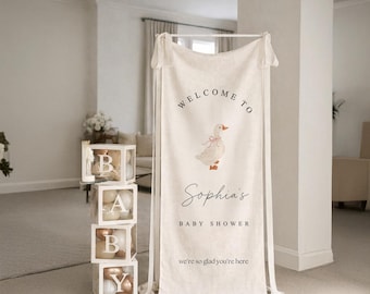 May include: A cream-colored fabric banner hanging from a white stand, featuring a goose illustration and the text "WELCOME TO Sophia's BABY SHOWER we're so glad you're here." Beside it are four stacked clear boxes containing balloons.