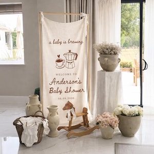 May include: A baby shower scene with a cream-colored banner that reads "a baby is brewing" and "Welcome to Anderson's Baby Shower 06.09.2026". The banner is framed by a wooden structure. Decorative vases, a rocking horse, and a basket with baby clothes are also present.