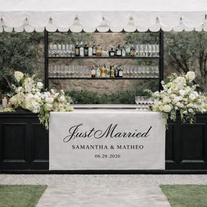 May include: A black bar with a white linen front featuring the words "Just Married", names, and a date. The bar is decorated with white floral arrangements and glassware. Shelves behind the bar hold bottles of liquor.