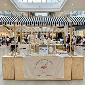 May include: A jewelry display booth with a black and white striped awning. The booth features necklaces, bracelets, and earrings. A banner displays the text "Home Grown Heart Restaurant & Bar" with a flamingo illustration.