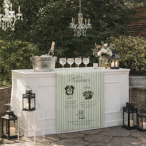 Peut inclure: Un bar ext&eacute;rieur blanc avec un chemin de table &agrave; rayures vertes et blanches affichant des illustrations de chiens et des noms de boissons. Le bar est &eacute;quip&eacute; de verres, d'une bouteille de champagne dans un seau &agrave; glace et de lanternes. Des lustres en cristal sont suspendus.