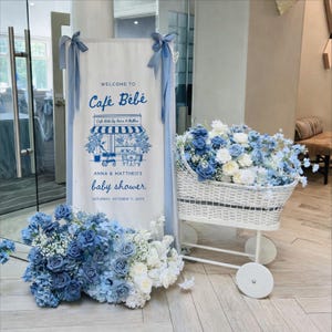 May include: A baby shower decoration featuring a sign that reads "Welcome to Café Bébé" with a blue and white illustration. A white wicker basket on wheels is filled with blue and white flowers, and more flowers are arranged at the base of the sign.