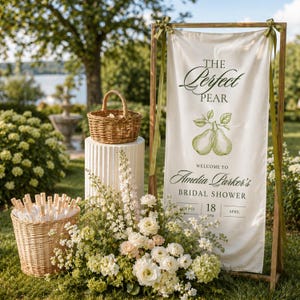 May include: A rustic wooden sign with the text "The Perfect Pear" and "Welcome to Amelia Parker's Bridal Shower" is displayed. A woven basket sits atop a white pedestal, and a basket of flowers and greenery are in the foreground.