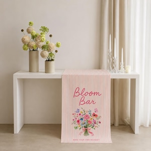 May include: A light pink and white vertical striped table runner featuring the text "Bloom Bar" in pink script above a colorful floral bouquet illustration and the text "MAKE YOUR OWN BOUQUET" in small pink capital letters at the bottom edge.