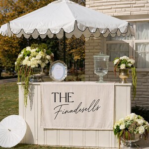 May include: A white outdoor bar with a scalloped-edge umbrella, featuring a beige fabric banner printed with the text THE Finadesells. The setup includes a glass beverage dispenser, a silver-toned sign, and three large floral arrangements.