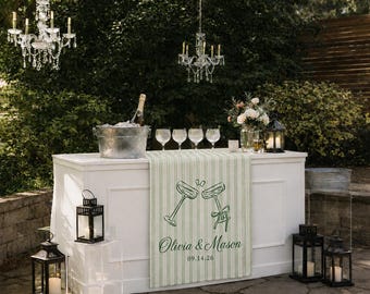 May include: A white outdoor bar with a green and white striped runner featuring champagne flutes and the names "Olivia & Mason" with a date. Includes champagne, glasses, lanterns, and chandeliers.