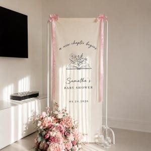 May include: A tall, off-white fabric banner suspended on a white metal frame, featuring the text 'a new chapter begins', a line-art illustration of an open book with flowers, 'Samatha's BABY SHOWER', and '01.24.2026', accented with pink satin ribbons.
