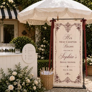 May include: A bridal shower banner with the text "Her Next Chapter Begins Welcome to Sophia's Bridal Shower May 24, 2026." The banner is cream-colored with burgundy accents and is displayed outdoors with a white umbrella.