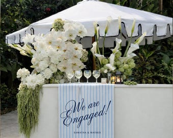 Puede incluir: Una barra al aire libre blanca con una pancarta a rayas azules y blancas que dice "We are Engaged!" con los nombres Samantha & Matheo. La barra está decorada con flores blancas, copas de vino y una sombrilla blanca.