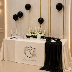 May include: A rectangular event table covered with a cream-colored tablecloth, featuring a central runner printed with a floral crest, the letter K, and the text Joshua & Beilay. The table includes black balloons, white candles, and flowers.