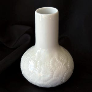 Könnte beinhalten: Eine weiße Keramikvase mit rundem Boden und zylindrischem Hals. Der Boden weist ein detailliertes, geprägtes Blumenmuster auf. Die Vase steht vor einem schwarzen Hintergrund, der ihre glatte, glänzende Oberfläche hervorhebt.