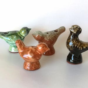 Könnte beinhalten: Vier Vogelpfeifen aus Keramik mit glänzender Oberfläche. Eine ist hellgrün, eine ist einfarbig rotbraun, eine ist gesprenkelt in Beige und Braun, und eine ist dunkelbraun mit einer strukturierten, gefleckten Optik. Jeder Vogel hat ein kleines Loch.