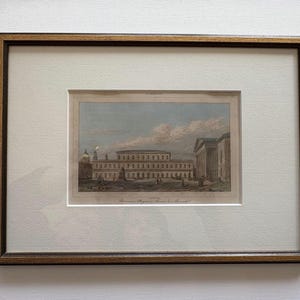 May include: Framed antique print of a European building with a detailed architectural design. The print is in a gold-toned wooden frame with a white mat. The artwork depicts a large building with columns and figures, under a cloudy sky.