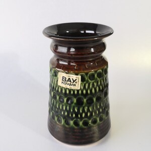 Puede incluir: Un jarrón de cerámica con borde y base marrón oscuro, que pasa a una sección media marrón más clara. El jarrón presenta un patrón texturizado de círculos verdes y el texto "BAY KERAMIK" en una etiqueta.