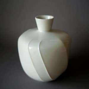 May include: A white ceramic vase with a unique, rounded shape and a small, cylindrical opening. The vase features a subtle, curved design element that wraps around its body, adding visual interest. The minimalist design is suitable for home decor.