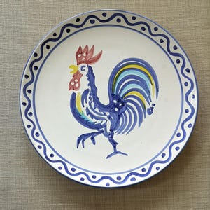 Puede incluir: Plato de cerámica blanco con un diseño de gallo pintado a mano en azul, amarillo y rojo. El plato tiene un borde azul ondulado con detalles punteados. Ideal para la decoración de la cocina o el comedor.