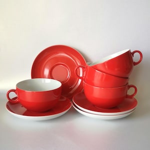 Pode incluir: Um conjunto de chávenas de chá e pires em cerâmica vermelha brilhante com um acabamento liso e lustroso. As chávenas apresentam uma forma de taça arredondada com uma pequena asa, enquanto os pires têm uma reentrância circular no centro.