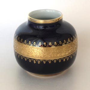 Può includere: Un piccolo vaso sferico in porcellana caratterizzato da una smaltatura blu navy profondo. Il centro del vaso è decorato con un'ampia fascia orizzontale in foglia d'oro metallizzato, accentuata da intricati motivi a filigrana sui bordi.