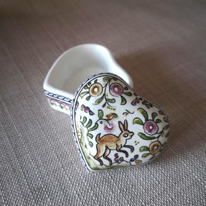 May include: A heart-shaped ceramic trinket box with a removable lid. The lid features a hand-painted design of a brown rabbit surrounded by stylised green leaves and small pink and yellow flowers. The base has a blue and white geometric border.