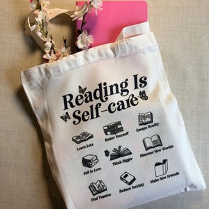 Op de afbeelding: Een witte draagtas met de tekst "Reading Is Self-care" en boekillustraties. De tas bevat een roze notitieboek en is versierd met roze en witte bloemen. Het ontwerp van de tas bevat de zinnen "Learn Lots" en "Make New Friends".
