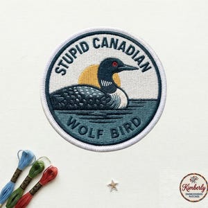 May include: Embroidered patch with a loon design, featuring the text "STUPID CANADIAN WOLF BIRD". The patch has a white border, a blue and green water scene, and a yellow sun in the background. The patch is round and has a textured appearance.