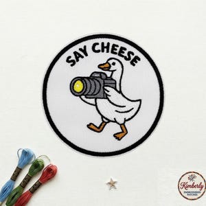 May include: Embroidered patch featuring a white goose holding a camera, with the text "SAY CHEESE" in a black circular border. The goose is walking with orange feet. The patch is on a white background.