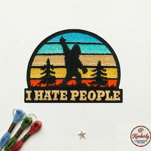 May include: Embroidered patch featuring a black silhouette of Bigfoot with an arm raised, set against a sunset design. The text "I HATE PEOPLE" is below the image. The patch has a black border and is on a white background.