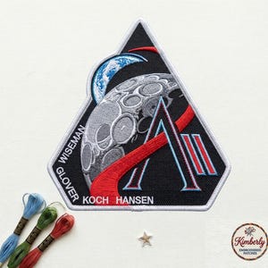 May include: Embroidered patch with a black background, featuring a stylized moon and Earth, with red and light blue accents. The patch includes the names WISEMAN, GLOVER, KOCH, and HANSEN. A white border surrounds the patch.