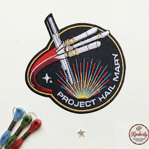 May include: Embroidered patch with a black background featuring a rocket design and the text "PROJECT HAIL MARY". The patch has a red and white trail, with the words "HAIL MARY" in white. A star and colorful rays are also included.