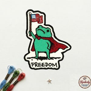 May include: Embroidered patch featuring a green frog wearing a red cape and holding an American flag. The word "FREEDOM" is below the frog. The patch has a black border and is on a white background.