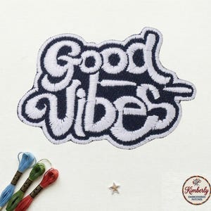 May include: Embroidered patch with the words "Good Vibes" in white script on a navy blue background. The patch has a white border and is surrounded by embroidery floss in blue, green, and red. A small star and a logo are also visible.