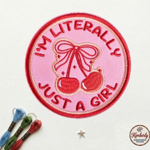 May include: Embroidered patch with a pink background and a red border. The patch features a red bow and cherries, with the text "I'M LITERALLY JUST A GIRL" in a circular design. The patch is likely for clothing or accessories.