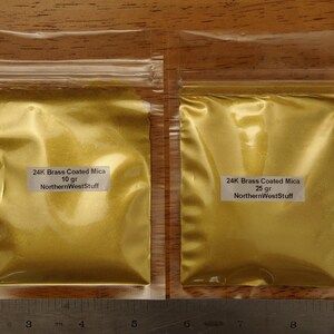 Puede incluir: Dos bolsas de pl&aacute;stico transparente llenas de polvo dorado. Cada bolsa tiene una etiqueta blanca con texto negro que dice "24K Brass Coated Mica" y "NorthernWestStuff". Una bolsa est&aacute; etiquetada "10 g" y la otra "25 g".