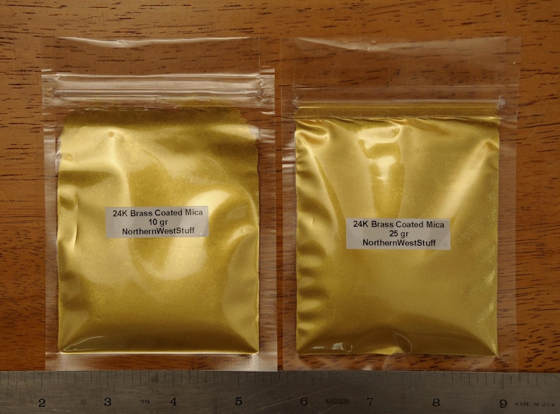 Puede incluir: Dos bolsas de pl&aacute;stico transparente llenas de polvo dorado. Cada bolsa tiene una etiqueta blanca con texto negro que dice "24K Brass Coated Mica" y "NorthernWestStuff". Una bolsa est&aacute; etiquetada "10 g" y la otra "25 g".