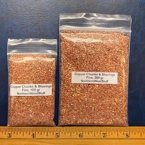 May include: Two clear plastic bags filled with copper chunks and shavings. The bags are labeled "Copper Chunks & Shavings Fine" with weights of 100 gr and 200 gr. The text "NorthernWestStuff" is also visible.