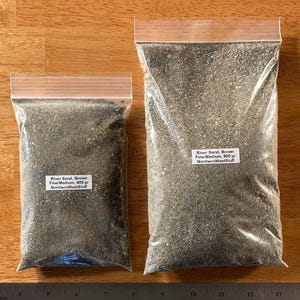 May include: Two clear plastic bags filled with brown river sand, labeled with text. The bags are on a wooden surface next to a metal ruler. The text on the bags reads "River Sand, Brown Fine/Medium, 400 gr NorthernWestStuff" and "River Sand, Brown Fine/Medium, 800 gr NorthernWestStuff".