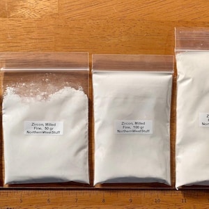 May include: Four clear plastic bags filled with white powder, labeled "Zircon, Milled Fine" with varying weights: 25 gr, 50 gr, 100 gr, and 200 gr. The bags are arranged in a row on a wooden surface.
