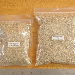 May include: Two clear plastic bags filled with fine vermiculite granules. The bags are labeled with the text "Fine Vermiculite" and "NorthernWest Stuff", with one bag containing 25 grams and the other 50 grams. The bags are on a wooden surface.