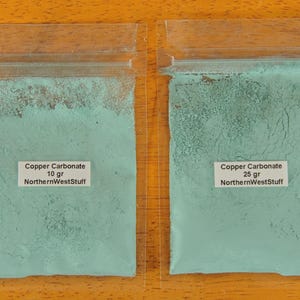 May include: Two clear plastic bags filled with a light blue powder, labeled "Copper Carbonate." One bag contains 10 grams, the other 25 grams. The text "NorthernWestStuff" is printed on the labels. The bags are on a wooden surface.