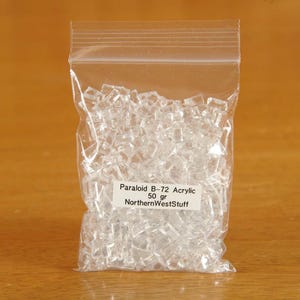 May include: A clear plastic bag filled with small, clear acrylic pieces. The bag has a label that reads "Paraloid B-72 Acrylic 50 gr NorthernWestStuff". The bag is sealed and sits on a wooden surface.