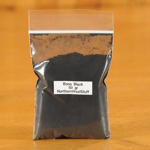 May include: A clear plastic bag filled with black powder, labeled "Bone Black 50 gr NorthernWestStuff." The bag is sealed at the top and sits on a wooden surface. The powder appears finely ground.