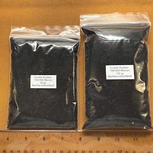 May include: Two clear plastic bags filled with black crumb rubber, each with a white label. The labels read "Crumb Rubber 150-500 Micron" and "NorthernWestStuff". One bag contains 50 grams, the other 100 grams. The bags are on a tan surface.