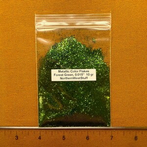 May include: A clear plastic bag filled with forest green metallic glitter flakes. The label reads "Metallic Color Flakes Forest Green, 0.015" 10 gr NorthernWestStuff". The bag is on a yellow-orange background.