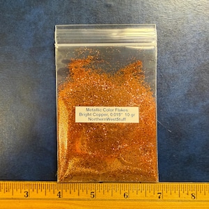 May include: A clear plastic bag filled with bright copper metallic flakes. The label reads "Metallic Color Flakes Bright Copper, 0.015" 10 gr NorthernWestStuff". A ruler is at the bottom of the image.