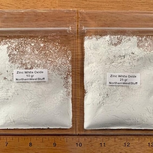 May include: Two clear plastic bags filled with white powder, labeled "Zinc White Oxide" with weights of 10 grams and 25 grams. The text "NorthernWestStuff" is also printed on the labels. The bags are placed on a wooden surface.