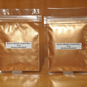 May include: Two clear plastic bags filled with bronze metallic coated mica powder. The bag on the left contains 10 grams, and the bag on the right contains 25 grams. Each bag has a white label with product information and the seller's name.