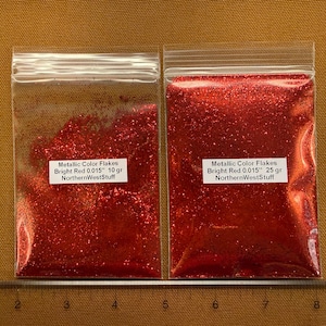 May include: Two clear plastic bags filled with bright red metallic glitter flakes. Each bag has a white label with black text. The bags are placed on a brown surface next to a ruler.