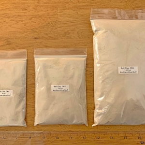 May include: Three clear plastic bags filled with off-white ball clay. The bags are labeled with the text "Ball Clay, OM4" and the weight: 50 grams, 100 grams, and 400 grams. The brand name "NorthernWestStuff" is also printed on the labels. A wooden ruler is at the bottom.