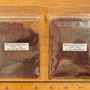 May include: Two clear plastic bags filled with blue, gold, and red glitter flakes. Each bag has a white label with the text "Blue Gold Red Flakes 0.015"" and "NorthernWestStuff". One bag contains 10 grams, the other 25 grams.